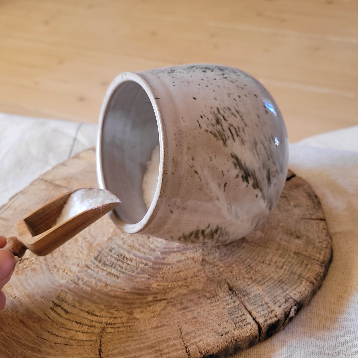 Gray ceramic salt pig with a speckled glaze, handmade by JogitaArtStudio, accompanied by a small wooden serving spoon.