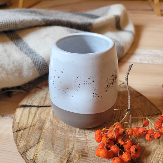 Alt Text: A handmade ceramic tumbler with a speckled white glaze and a gray base. The tumbler has dimples along its sides and is partially covered by a cozy blanket. There are also a few red berries scattered nearby.
