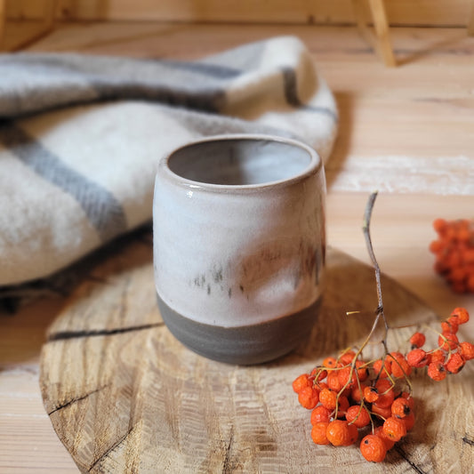 A handmade ceramic tumbler with a gray base and a white glaze. The tumbler has a textured surface with subtle lines and imperfections. It's resting on a wooden surface with a small branch and some red berries nearby.