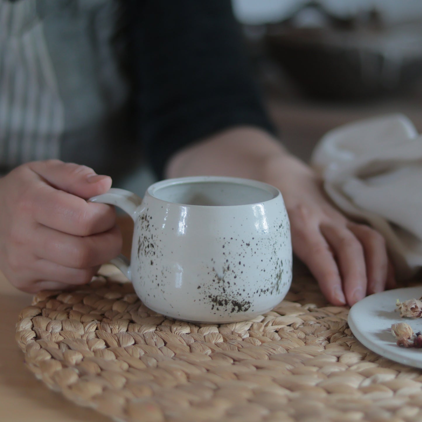 Hand houdt het handvat van een witte gespikkelde cappuccino mok van aardewerk vast op een geweven placemat, met op de achtergrond een schaaltje gedroogde bloemen