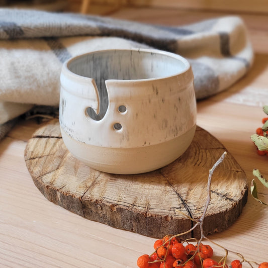 Handmade ceramic yarn bowl with a textured glaze, resting on a wooden board. A skein of yarn is partially visible in the bowl, and there are red berries scattered around.