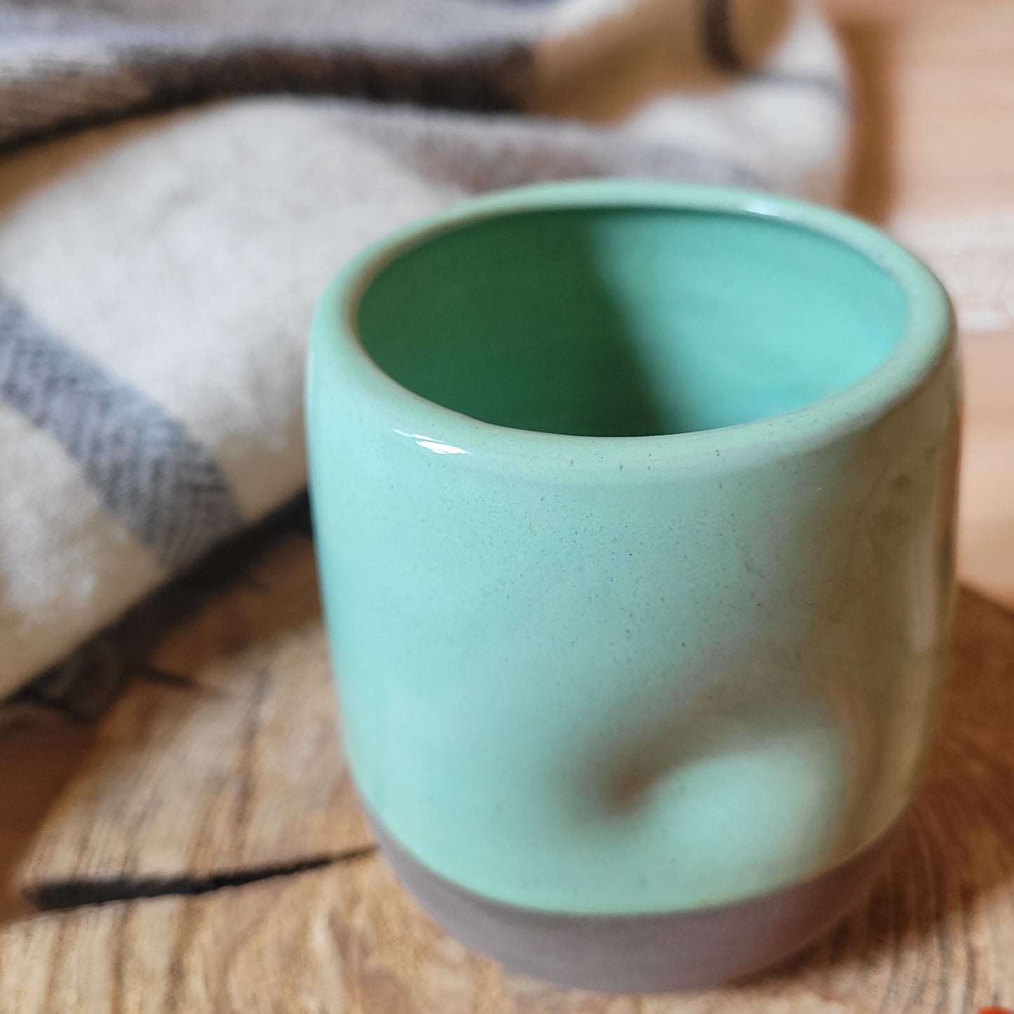 A mint green ceramic tumbler with a gray base. The tumbler has a textured surface and is partially covered by a cozy blanket. There are a few red berries scattered on a wooden surface nearby.