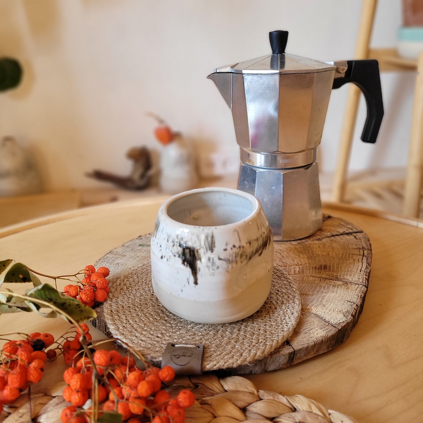 A modern white ceramic double espresso mug with a speckled glaze, placed on a wooden stand, with a cappuccino machine and decorative oranges in the background.