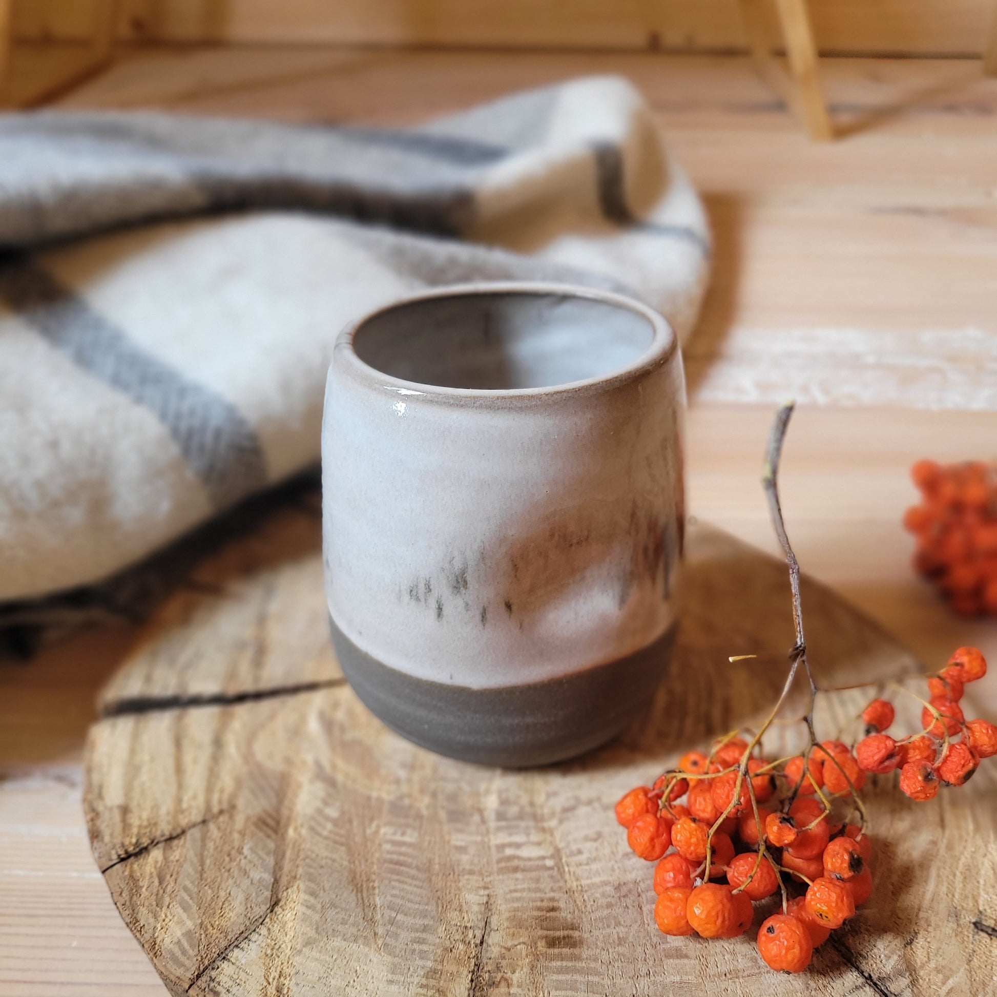  A handmade ceramic tumbler with a gray base and a white glaze. The tumbler has a textured surface with subtle lines and imperfections. It's resting on a wooden surface with a small branch and some red berries nearby.