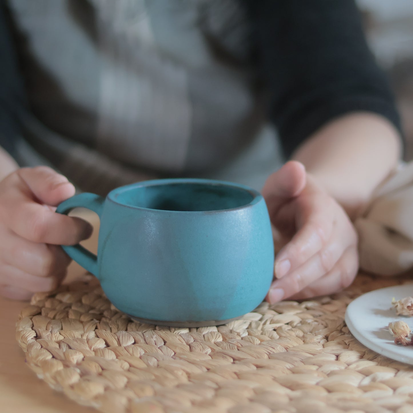 Hand holding a teal matte stoneware cappuccino mug with soft blue-green colour transitions, on a woven placemat with a small plate of dried flowers in the background