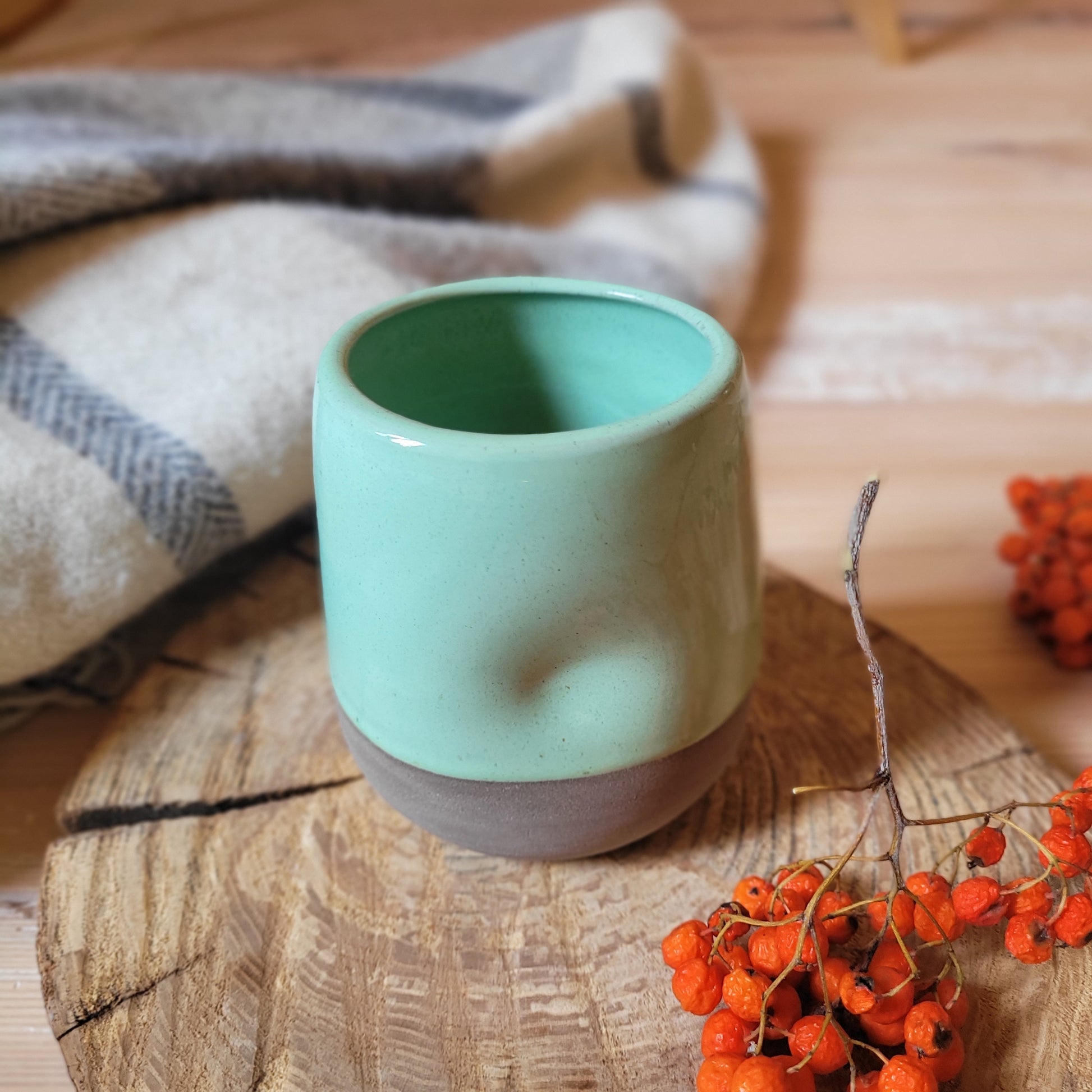 A mint green ceramic tumbler with a gray base. The tumbler has a textured surface and is partially covered by a cozy blanket. There are also a few red berries scattered on a wooden surface nearby.